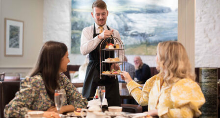 A waiter serves afternoon tea to two women in a cosy, elegantly decorated hotel restaurant.
