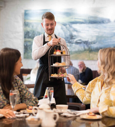 Two women enjoy afternoon tea at a cosy hotel, served by a friendly waiter.