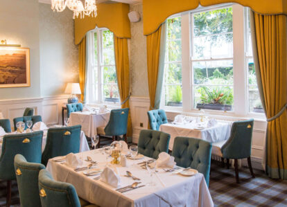 Elegant dining room with set tables and teal chairs at Old Ground Hotel. Sunlight streams through large windows.