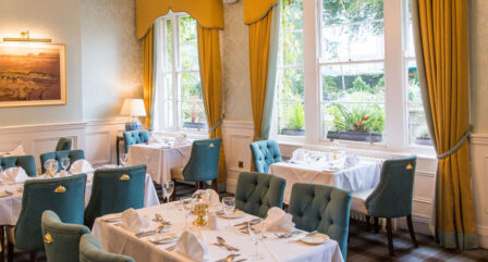 Elegant dining room with set tables and teal chairs at Old Ground Hotel. Sunlight streams through large windows.