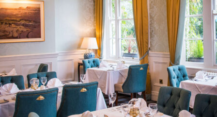 Elegant hotel dining room with blue chairs, white tablecloths, and natural light from tall windows.