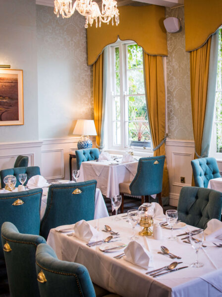 Elegant hotel dining room with blue chairs, white tablecloths, and natural light from tall windows.