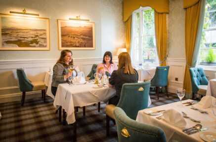 Three friends enjoy dining in a cosy, elegant hotel restaurant with warm decor and large windows.