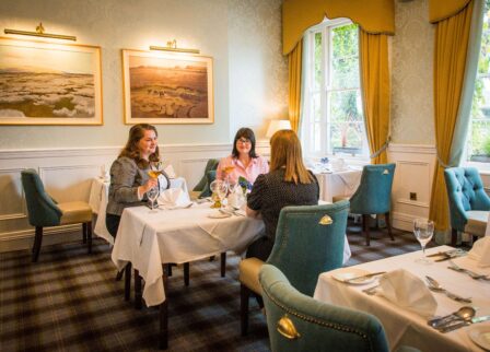 Three friends enjoy dining in a cosy, elegant hotel restaurant with warm decor and large windows.