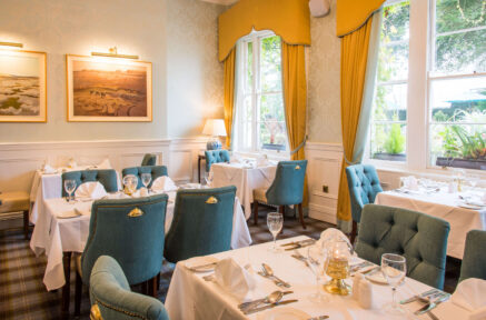 Elegant dining room with blue chairs, set tables, and warm sunlight through large windows.
