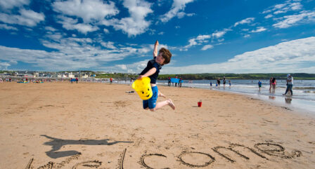 Young child joyfully jumps on sandy beach with "Welcome" written in the sand; families in background.