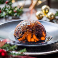 Festive Christmas pudding aflame on elegant plate, surrounded by holiday decor.