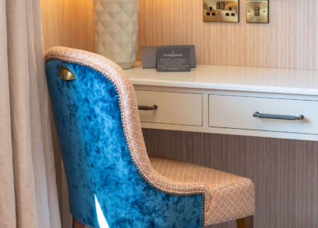 Cosy hotel room with a velvet blue chair, a stylish lamp, and a welcoming atmosphere.
