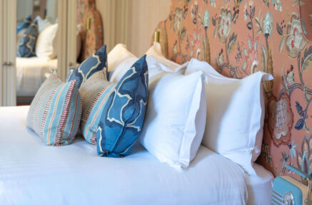 Elegant hotel room with white and blue pillows on a neatly made bed, floral-patterned headboard.