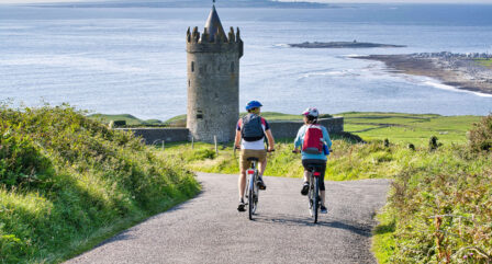 Couple cycling towards a historic tower by the scenic coast, enjoying a sunny day.