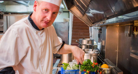Chef preparing roasted meat in hotel kitchen with fresh vegetables. Warm, inviting atmosphere.