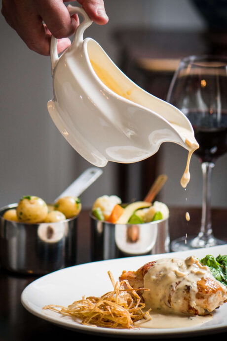 Creamy sauce being poured over a gourmet dish beside a glass of red wine at a hotel restaurant.