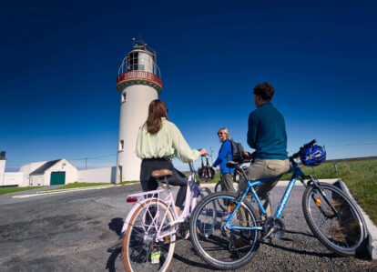 A couple and a friend enjoy a sunny bike ride by a historic lighthouse, engaged in conversation.