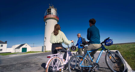 A couple and a friend enjoy a sunny bike ride by a historic lighthouse, engaged in conversation.