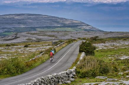 A couple cycling on a scenic, rural road with lush hills and expansive landscapes.