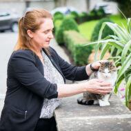 A woman gently strokes a cat on a stone wall in a lush garden outside a hotel.
