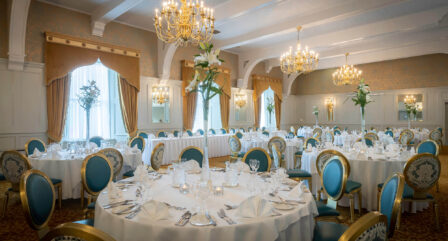 Elegant hotel dining room with round tables, set for a banquet, featuring chandeliers and floral decor.