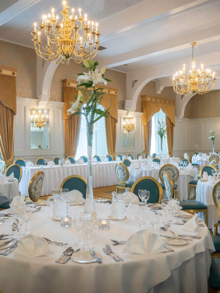 Elegant hotel dining room with round tables, set for a banquet, featuring chandeliers and floral decor.