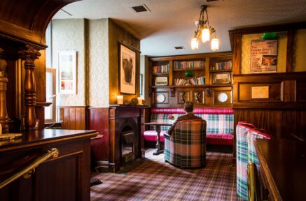 Cosy hotel lounge with a person relaxing by a vintage fireplace, surrounded by warm plaid seating and bookshelves.
