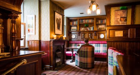 Cosy hotel lounge with a person relaxing by a vintage fireplace, surrounded by warm plaid seating and bookshelves.