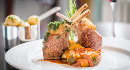 Elegant lamb chops with vegetables on a white plate, alongside seasoned potatoes and red wine.