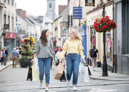 Two women smiling and shopping on a lively street with hanging flowers and quaint shops.