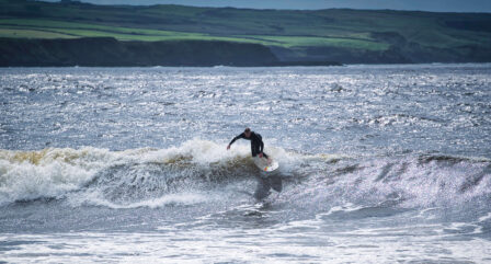 A surfer catching a wave on a sunny day, with scenic cliffs in the background.