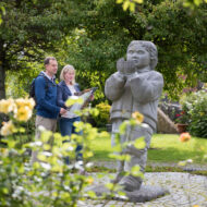Couple exploring lush hotel garden, admiring statue surrounded by blooming flowers.