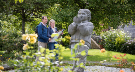 Couple exploring lush hotel garden, admiring statue surrounded by blooming flowers.