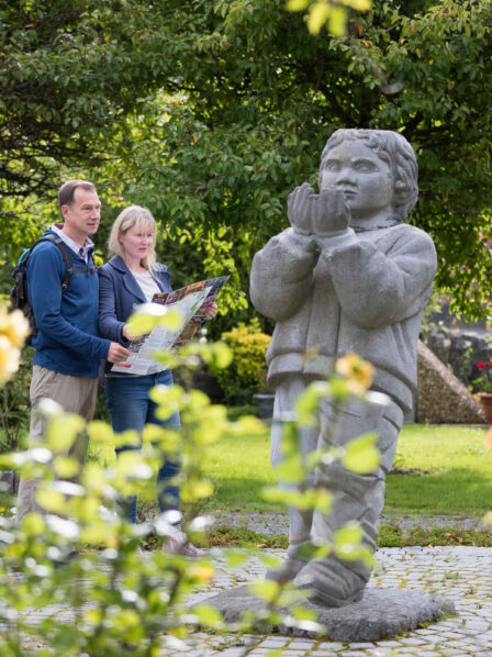 Couple exploring lush hotel garden, admiring statue surrounded by blooming flowers.