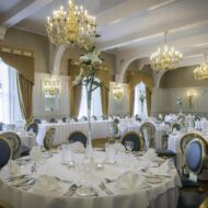 Elegant hotel dining room set for a formal event, with round tables, chandeliers, and floral centrepieces.