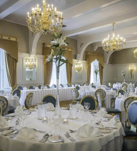 Elegant hotel dining room set for a formal event, with round tables, chandeliers, and floral centrepieces.