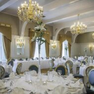Elegant dining room with round tables set for guests, featuring chandeliers and tall floral arrangements.