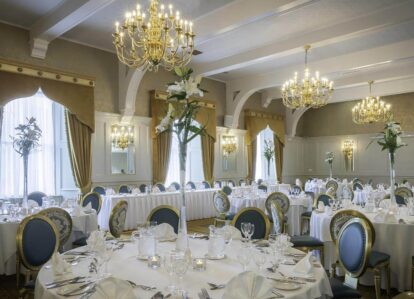 Elegant dining room with round tables set for guests, featuring chandeliers and tall floral arrangements.