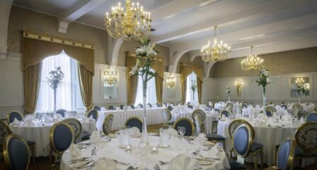 Elegant dining room with round tables set for guests, featuring chandeliers and tall floral arrangements.