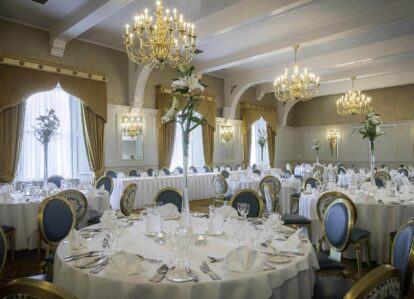 Elegant dining room with round tables set for an event, featuring chandeliers and floral centrepieces.