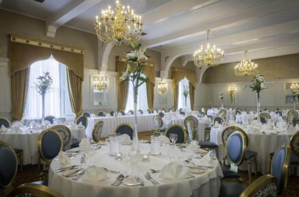 Elegant dining room with round tables set for an event, featuring chandeliers and floral centrepieces.