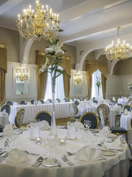 Elegant dining room with round tables set for an event, featuring chandeliers and floral centrepieces.