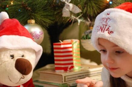 Young girl in Santa hat, with teddy bear, enjoying festive moments under a decorated Christmas tree.