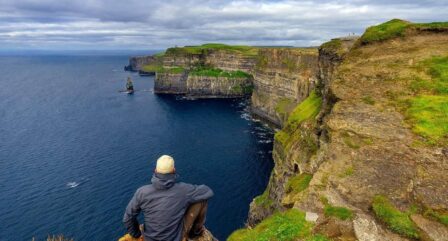 Person enjoying scenic view of Cliffs of Moher, with lush greenery and dramatic coastal cliffs.