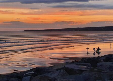 Serene beach sunset with surfers enjoying the waves, reflecting warm orange hues.