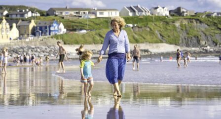 Smiling woman and child walking on a lively beach, with colourful houses and green cliffs in the background.