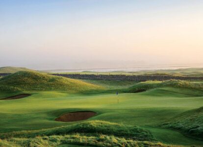 A serene golf course at sunrise, surrounded by rolling green hills near a distant stone ruin.