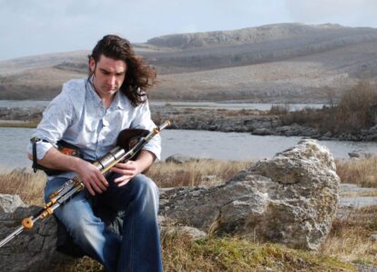 A man plays the uilleann pipes by a serene lake, surrounded by rugged hills and rocks.