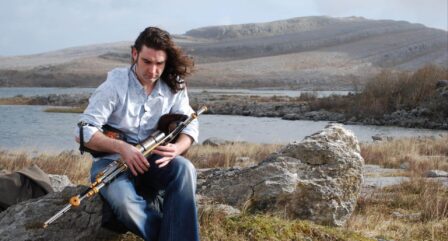 A man plays the uilleann pipes by a serene lake, surrounded by rugged hills and rocks.