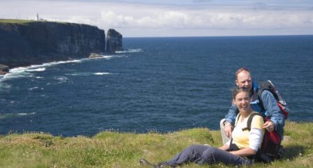 A couple relaxes on a grassy cliffside overlooking the ocean, with a lighthouse in the distance.