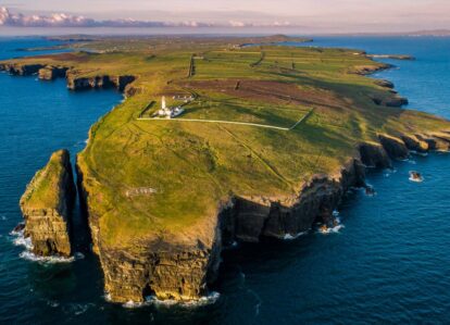 Aerial view of scenic coastal cliffs with lush greenery and a lighthouse overlooking the ocean.