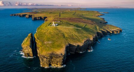 Aerial view of scenic coastal cliffs with lush greenery and a lighthouse overlooking the ocean.