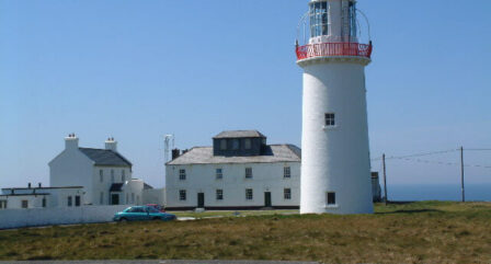 Historic lighthouse with nearby white buildings on a sunny day by the sea.