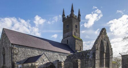 Historic stone church with tall spires under a blue sky, surrounded by lush greenery.